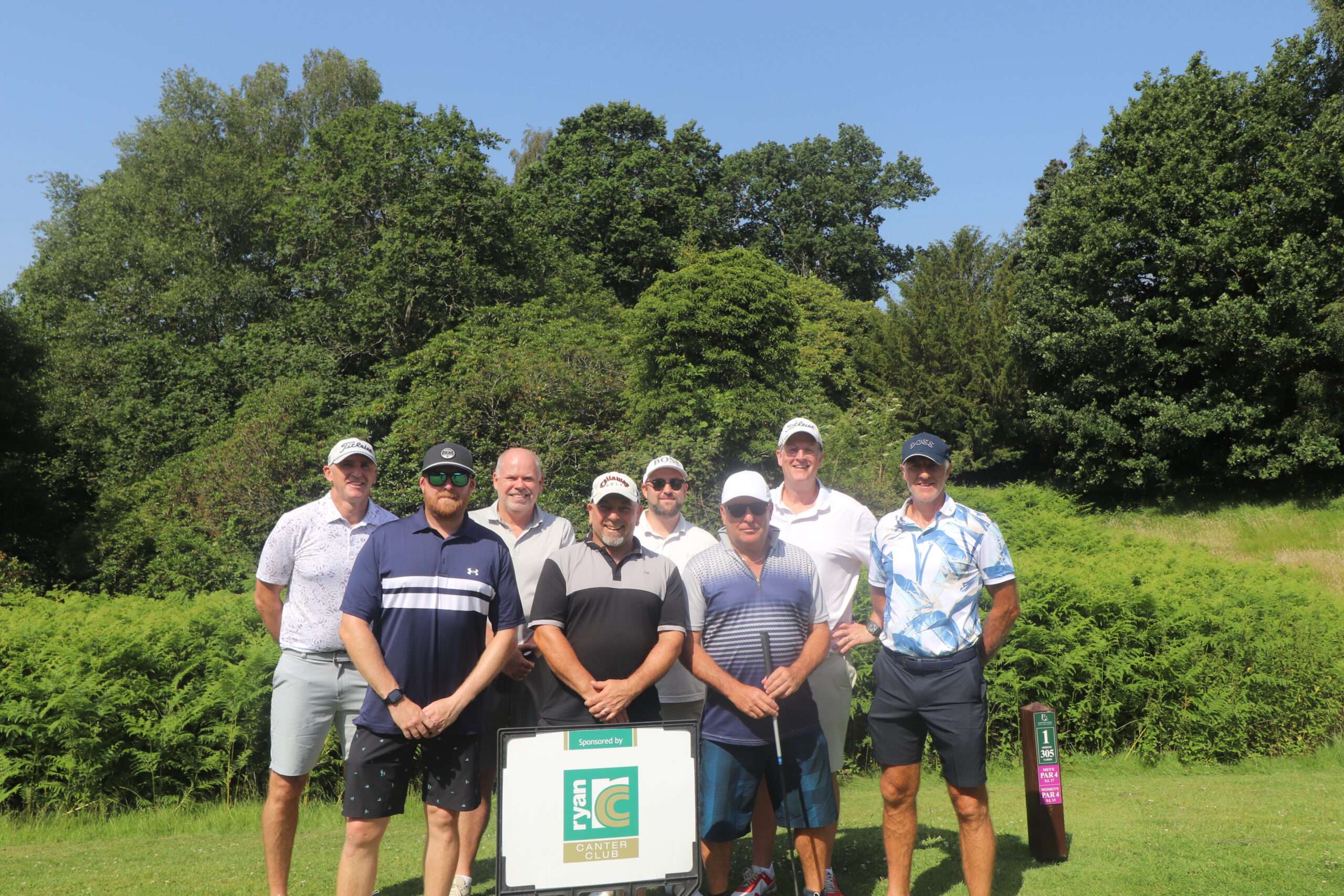 A group of men in golfing attire stand on grass behind a sign advertising Ryan Canter Club. There is bracken and trees in the background.