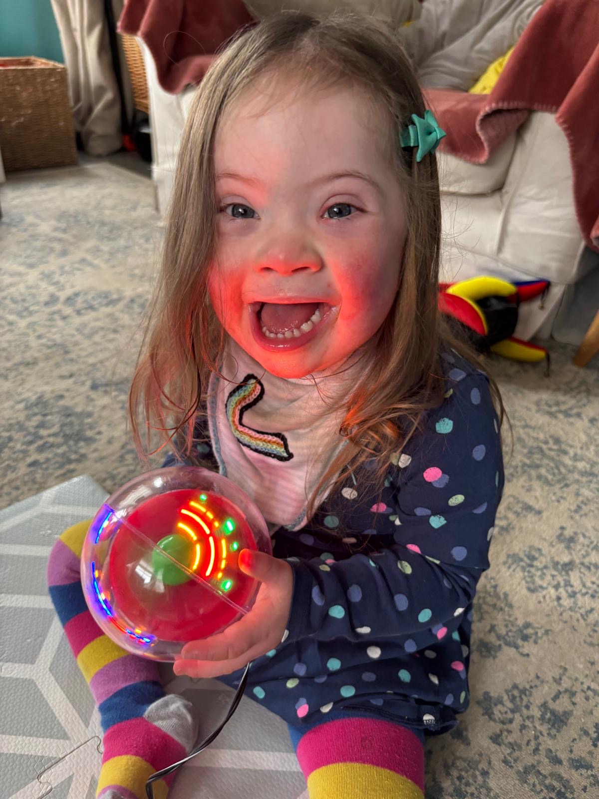 a young girl with down syndrome holds a light up ball flashing in lots of different colours. She is wearing a blue onesie with colourful polka dots and colourful socks.