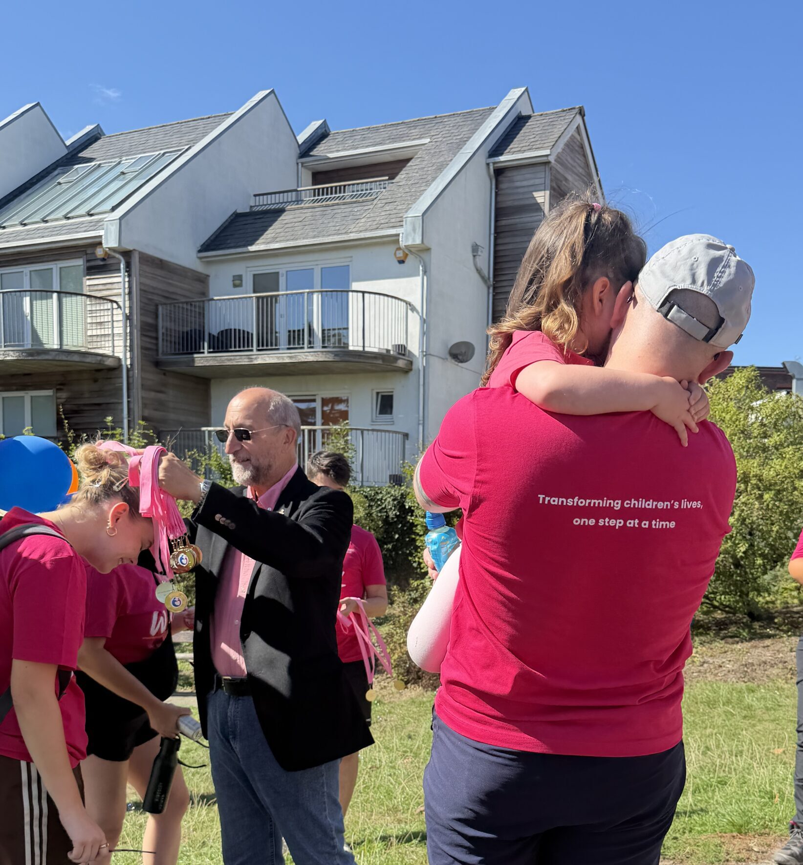 A man in a pink t shirt is holding a little girl in his arms. Behind them, walkers are being given medals for completing a sponsored walk.