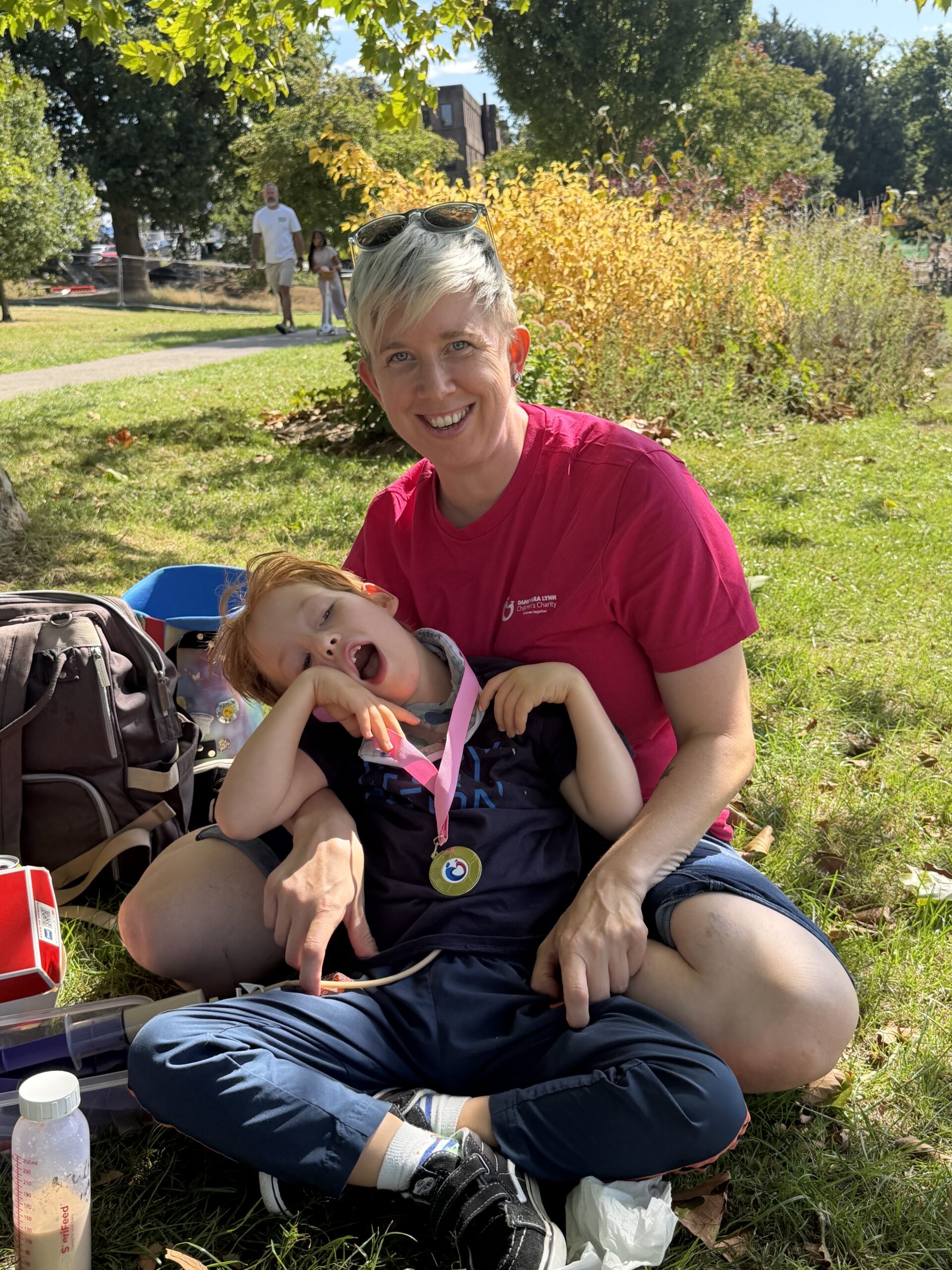A young boy with cerebral palsy sits on his mums lap. He has a medal around his neck from a sponsored walk. His mum is wearing a pink t shirt and sunglasses on her head.
